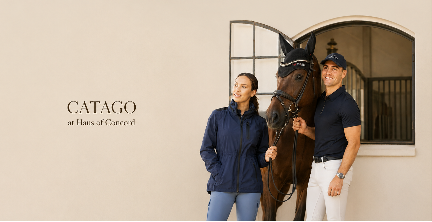 A woman and a man stand beside a brown horse in front of a beige wall with the text 'CATAGO at Haus of Concord.' The man wears a navy polo shirt, white riding pants, and a dark cap, while the woman wears a navy jacket and light blue pants.