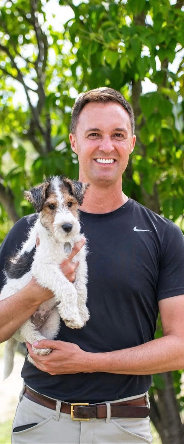 A smiling man in a black Nike t-shirt and khaki pants holds a small black and white dog outdoors in front of a leafy green tree.