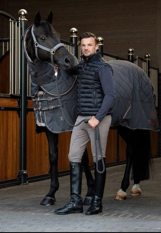 A man in equestrian attire stands with a black horse in a stable, wearing a dark quilted puffer vest, beige breeches, and black riding boots. The horse is covered with a dark gray quilted blanket.