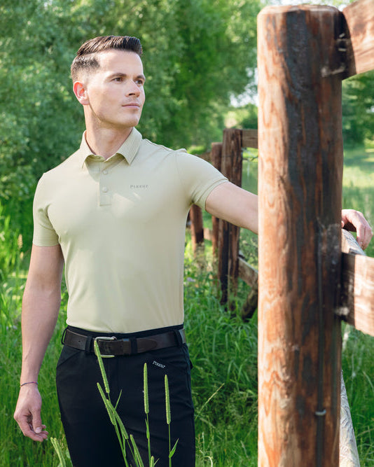 A man wearing a light beige short-sleeve polo shirt and black pants stands beside a rustic wooden fence in a grassy outdoor field.