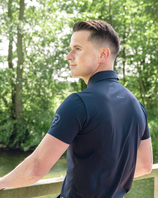 A man in a dark navy blue short-sleeve athletic shirt stands on a wooden bridge by a river, looking out toward a wooded area.